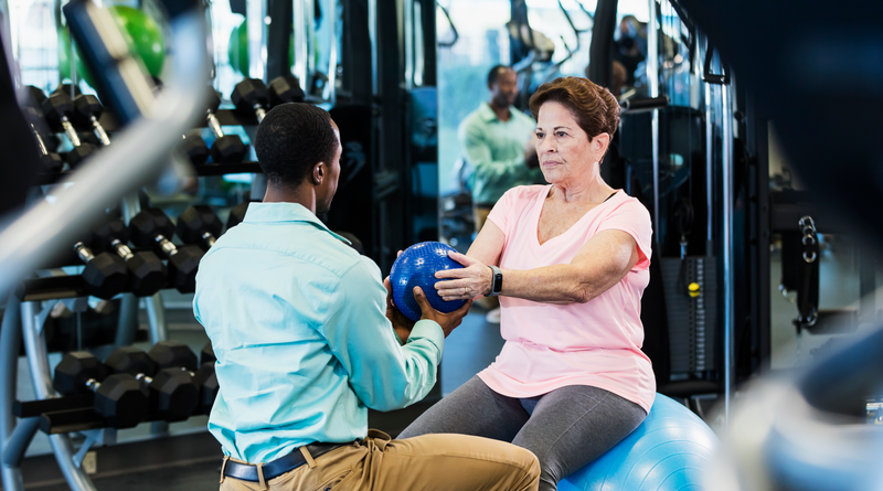 Senior woman doing physical therapy with medicine ball - stock photo
An African-American man in his 30s working as a physical therapist helping a senior woman in her 60s recover from an injury, doing strengthening exercises in a gym. She is sitting on an exercise ball and he is kneeling in front of her. They are passing a medicine ball back and forth.